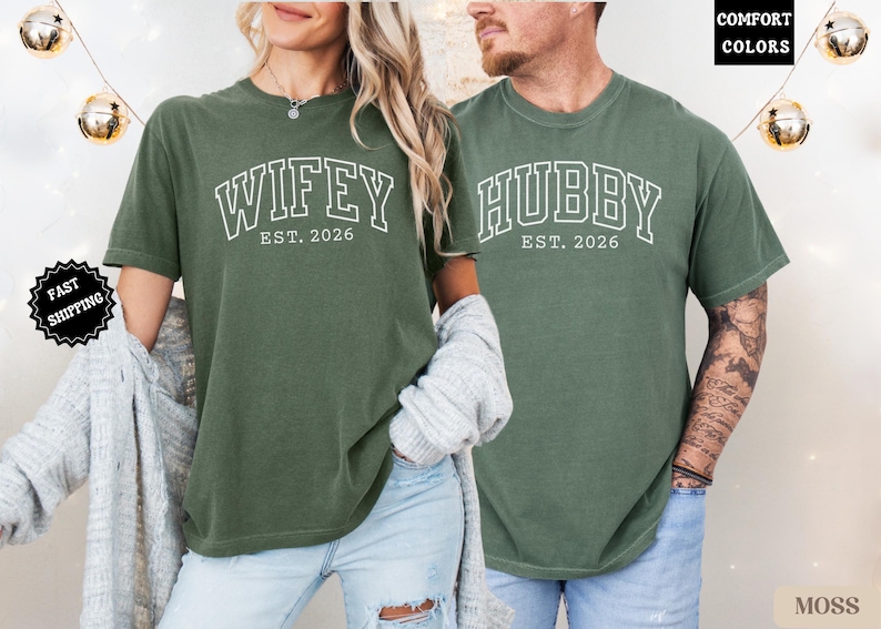 May include: Two moss green t-shirts. One says "Wifey Est. 2026" in white block letters, the other says "Hubby Est. 2026". The shirts are worn by a couple. The background is neutral with decorative lights.