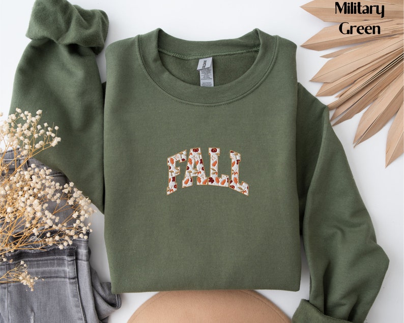 May include: A green sweatshirt with the word "FALL" printed on the front in a white font with a brown and orange floral pattern.