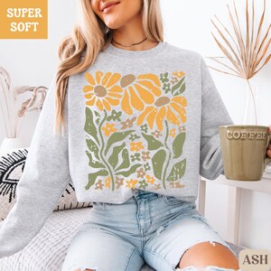 May include: A light gray sweatshirt with a floral design. The design features large yellow flowers, smaller orange and yellow flowers, and green leaves. The words "SUPER SOFT" are in a yellow box in the upper left corner. The word "ASH" is in a small box in the lower right corner.