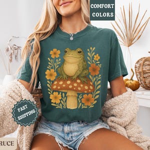 May include: A teal blue t-shirt featuring a graphic of a green frog sitting on a mushroom with yellow and orange flowers. The text "COMFORT COLORS" is on a white tag attached to the shirt.
