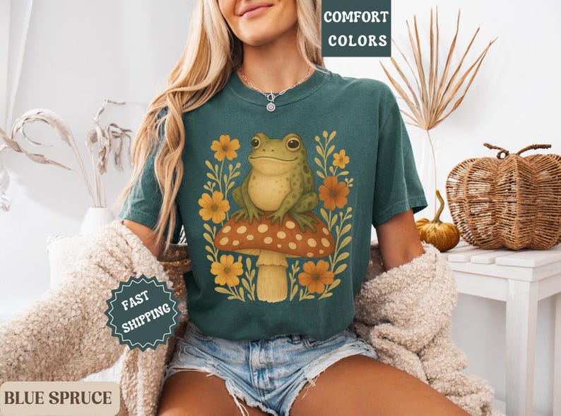 May include: A teal blue t-shirt featuring a graphic of a green frog sitting on a mushroom with yellow and orange flowers. The text "COMFORT COLORS" is on a white tag attached to the shirt.