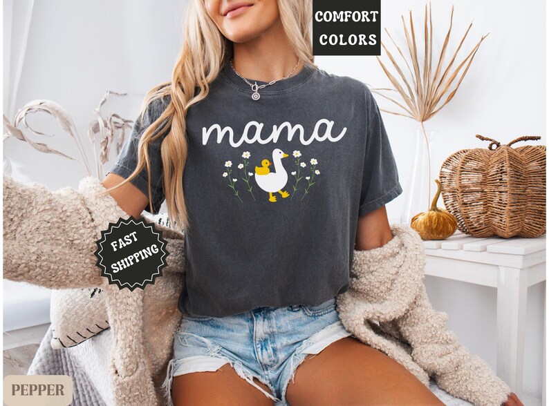May include: A dark gray t-shirt with the word "mama" printed in white. A white duck with a yellow chick is printed on the shirt with a floral design around it.