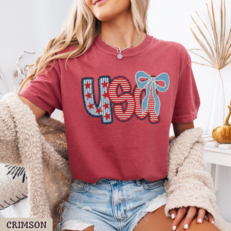 Op de afbeelding: Karmizijnrood T-shirt met een patriottisch "USA" ontwerp. De letters zijn versierd met rood-witte strepen en blauwe sterren, en een blauwe strik. Het shirt wordt gedragen met een spijkerbroek.
