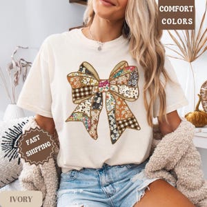 May include: Ivory t-shirt with a large bow design made from various patterned fabrics. The bow incorporates floral, plaid, and solid patches in brown, pink, and blue. The words "COMFORT COLORS" are printed on the top right.