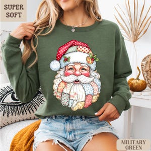 May include: Olive green sweatshirt featuring a colorful Santa Claus graphic. The Santa design has a patchwork beard and hat with red polka dots. The words "SUPER SOFT" are in the upper left corner, and "MILITARY GREEN" is in the lower right corner.