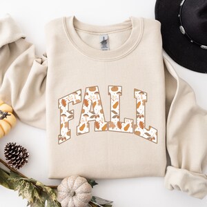 K&ouml;nnte beinhalten: Ein beiger Pullover mit dem Wort "FALL" in gro&szlig;en Buchstaben bedruckt. Die Buchstaben sind mit einem Muster aus Herbstbl&auml;ttern, Eicheln und K&uuml;rbissen gef&uuml;llt.