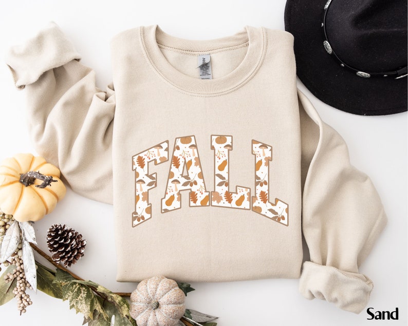 K&ouml;nnte beinhalten: Ein beiger Pullover mit dem Wort "FALL" in gro&szlig;en Buchstaben bedruckt. Die Buchstaben sind mit einem Muster aus Herbstbl&auml;ttern, Eicheln und K&uuml;rbissen gef&uuml;llt.