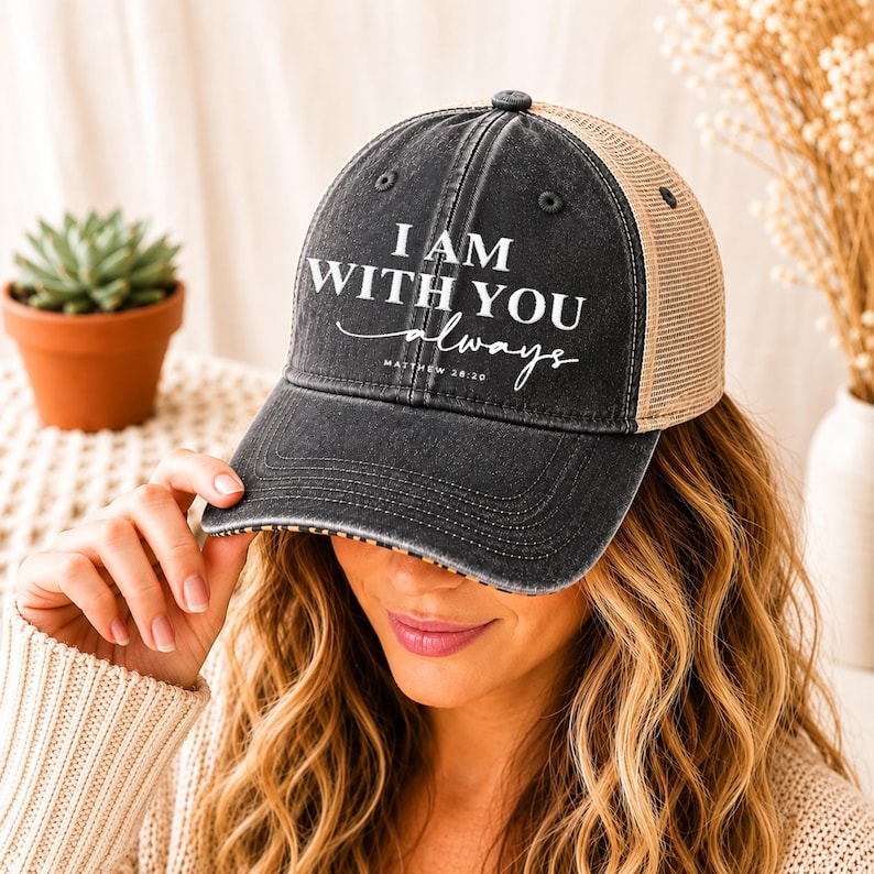 May include: A black and tan trucker hat with the text "I AM WITH YOU always" in white script. The hat has a curved brim and a mesh back. The hat is a casual accessory.