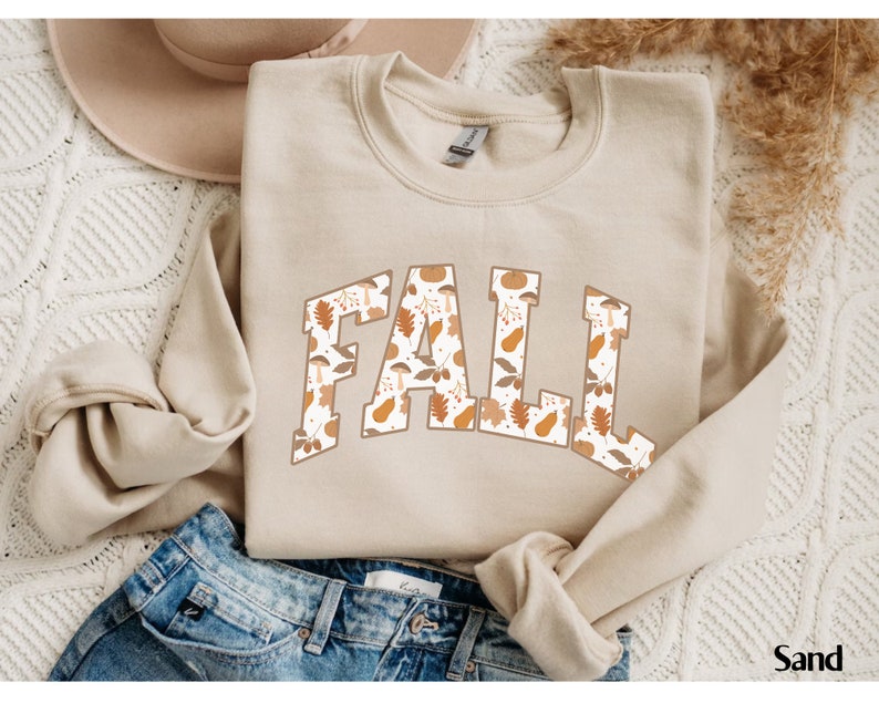 May include: Sand-colored sweatshirt with the word "FALL" in large, block letters. The letters are filled with a pattern of fall leaves, mushrooms, and other autumn-themed designs. The sweatshirt is folded on top of blue jeans.