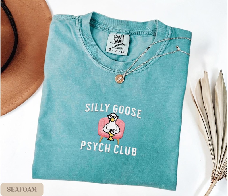 May include: Seafoam green t-shirt with a white embroidered design of a goose wearing glasses and sitting in a pink armchair. The text "Silly Goose Psych Club" is embroidered below the goose.