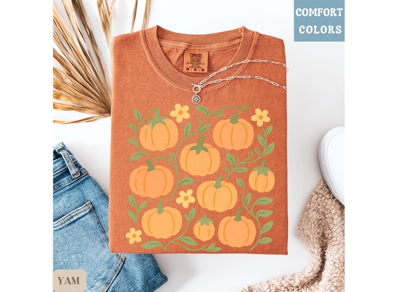 May include: Orange t-shirt with a pumpkin graphic. The shirt features a design of orange pumpkins, green vines, and yellow flowers. The words "COMFORT COLORS" are printed in the upper right corner. A silver necklace is on top of the shirt.