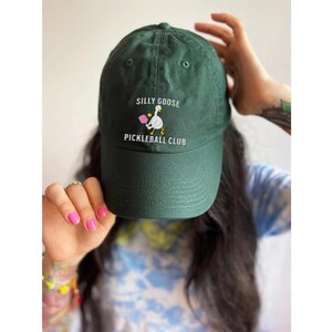 May include: A dark green baseball cap with the text "Silly Goose Pickleball Club" and a white goose holding a pink paddle embroidered on the front.