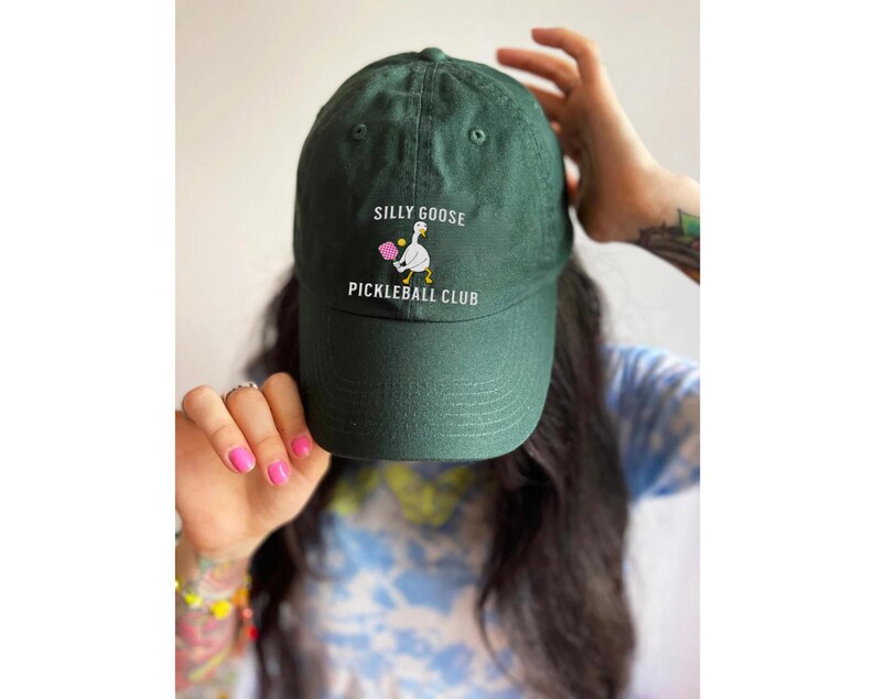 May include: A dark green baseball cap with the text "Silly Goose Pickleball Club" and a white goose holding a pink paddle embroidered on the front.