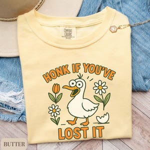 May include: Butter-colored t-shirt with a cartoon duck graphic. The duck has a surprised expression and is surrounded by flowers and the text "HONK IF YOU'VE LOST IT". The shirt is on a denim background.