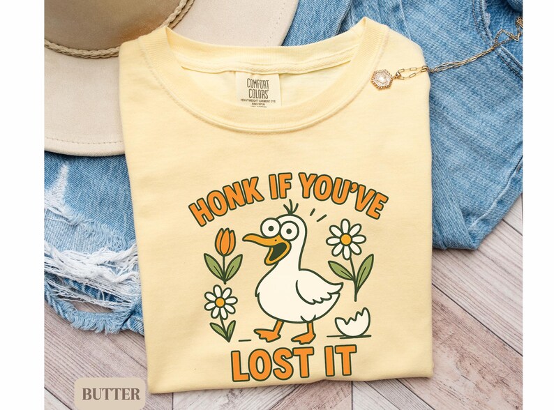 May include: Butter-colored t-shirt with a cartoon duck graphic. The duck has a surprised expression and is surrounded by flowers and the text "HONK IF YOU'VE LOST IT". The shirt is on a denim background.