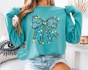 May include: Seafoam green long-sleeve sweatshirt featuring a Christmas bow design made of colorful string lights. The text "COMFORT COLORS" is in a tan box in the upper right corner. The text "SEA FOAM" is in a box at the bottom left.