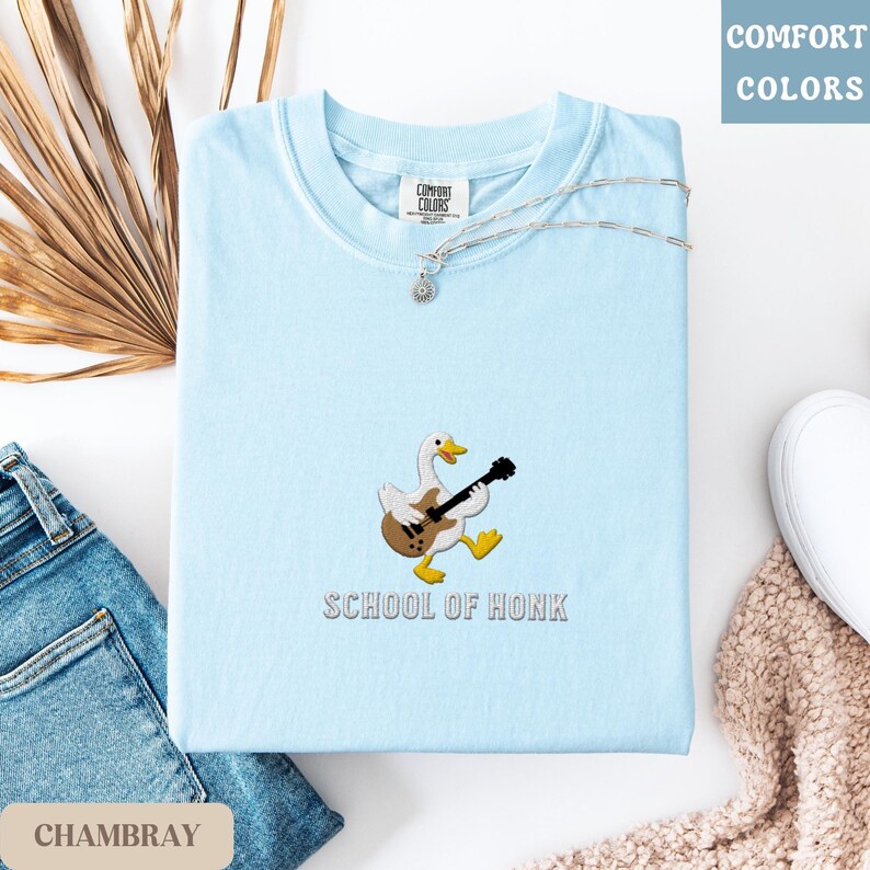 May include: A light blue t-shirt with a cartoon duck playing a guitar, with the text "SCHOOL OF HONK" below. The shirt is folded, with a silver necklace and a pair of jeans visible. The words "COMFORT COLORS" are in the top right corner.