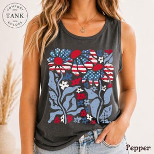 Op de afbeelding: Een donkergrijze tanktop met een bloemenontwerp met rode, witte en blauwe accenten. De bloemen hebben een patroon dat lijkt op de Amerikaanse vlag. De tanktop heeft de woorden "Comfort Tank Colors" en het woord "Pepper".