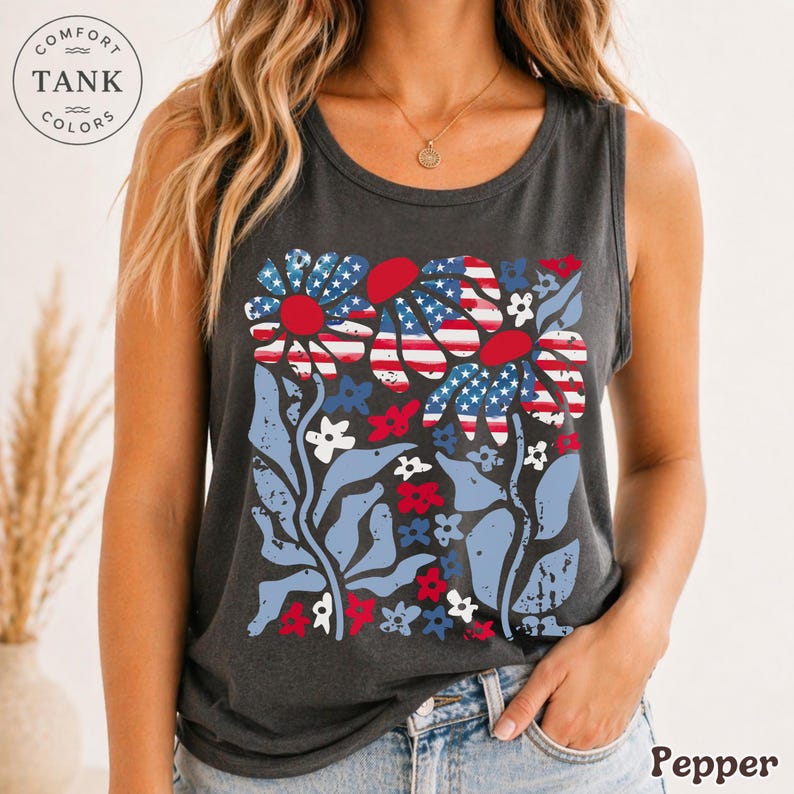 Op de afbeelding: Een donkergrijze tanktop met een bloemenontwerp met rode, witte en blauwe accenten. De bloemen hebben een patroon dat lijkt op de Amerikaanse vlag. De tanktop heeft de woorden "Comfort Tank Colors" en het woord "Pepper".