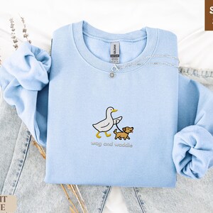 May include: Light blue sweatshirt with a crew neck. The sweatshirt features an embroidered design of a white duck walking a brown dog on a leash. The text "wag and waddle" is below the design. The words "SUPER SOFT" and "LIGHT BLUE" are also visible.
