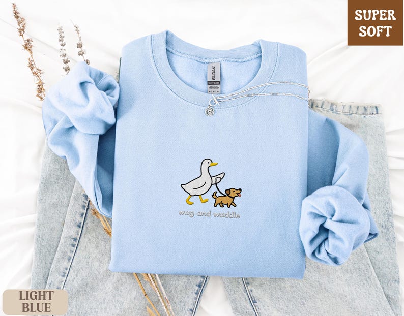 May include: Light blue sweatshirt with a crew neck. The sweatshirt features an embroidered design of a white duck walking a brown dog on a leash. The text "wag and waddle" is below the design. The words "SUPER SOFT" and "LIGHT BLUE" are also visible.
