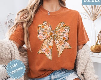 May include: Burnt orange t-shirt featuring a large floral bow design with gold glitter accents. The bow has a cream background with peach and orange flowers. The shirt has the words "COMFORT COLORS" in a blue box.