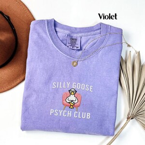 May include: A purple t-shirt with the text "Silly Goose Psych Club" and a cartoon goose wearing glasses sitting in a pink armchair.