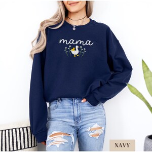 May include: A navy blue sweatshirt with the word "mama" embroidered on the front. The word is in a white cursive font and is surrounded by small white flowers. A white duck with a yellow chick is embroidered in the center of the flowers.