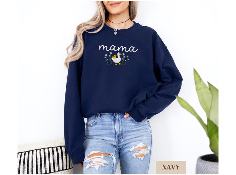 May include: A navy blue sweatshirt with the word "mama" embroidered on the front. The word is in a white cursive font and is surrounded by small white flowers. A white duck with a yellow chick is embroidered in the center of the flowers.