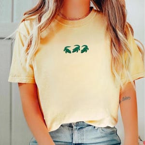 May include: A light yellow t-shirt with three green alligator designs. The shirt has short sleeves and a crew neck. The shirt is made of a soft material and is a casual style.