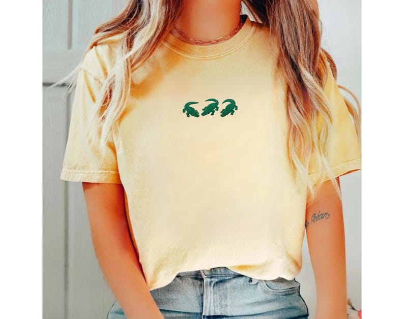 May include: A light yellow t-shirt with three green alligator designs. The shirt has short sleeves and a crew neck. The shirt is made of a soft material and is a casual style.