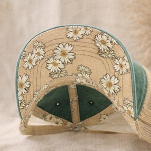 May include: A baseball cap with a floral daisy pattern on the underside of the brim. The cap features a green bill and a tan mesh back. The daisy pattern is white and yellow on a tan background.