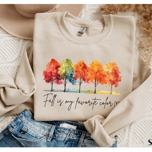 May include: A beige sweatshirt with a watercolor illustration of five trees with colorful leaves. The text "Fall is my favorite color" is written in black cursive below the trees.