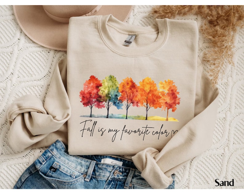 May include: A beige sweatshirt with a watercolor illustration of five trees with colorful leaves. The text "Fall is my favorite color" is written in black cursive below the trees.