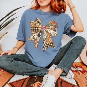 May include: A blue t-shirt featuring a large bow design made of various patterned fabrics. The bow has a patchwork appearance with floral, plaid, and solid sections. The shirt is paired with blue jeans.