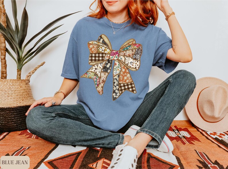 May include: A blue t-shirt featuring a large bow design made of various patterned fabrics. The bow has a patchwork appearance with floral, plaid, and solid sections. The shirt is paired with blue jeans.