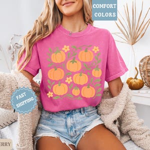 May include: A magenta t-shirt featuring a pumpkin design. The graphic includes orange pumpkins, green vines, and yellow flowers. The shirt is paired with denim shorts. The words "COMFORT COLORS" are in a light blue box.