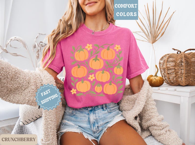 May include: A magenta t-shirt featuring a pumpkin design. The graphic includes orange pumpkins, green vines, and yellow flowers. The shirt is paired with denim shorts. The words "COMFORT COLORS" are in a light blue box.