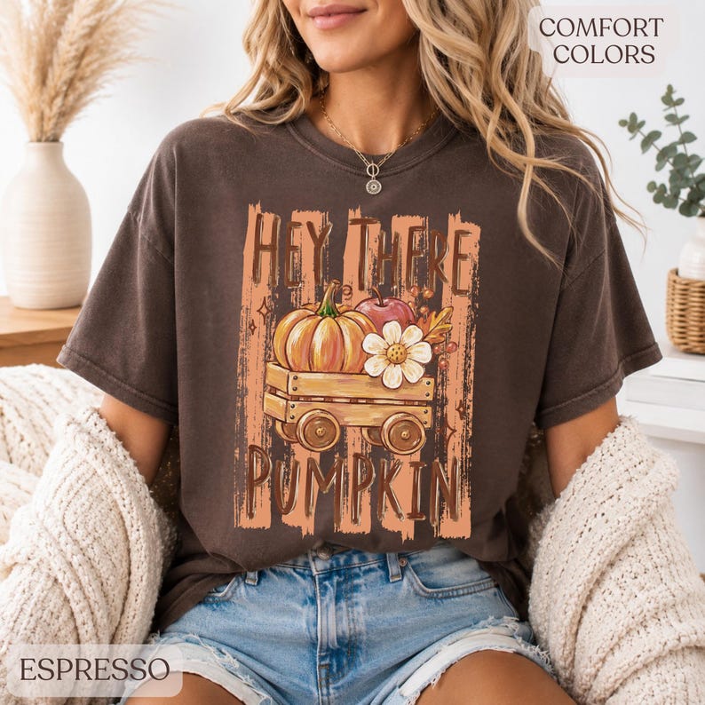 Op de afbeelding: Een espresso-kleurig t-shirt met korte mouwen en een grafische print van een houten wagen met een pompoen, een appel en een witte bloem. De tekst "HEY THERE PUMPKIN" is in een verweerd lettertype boven en onder de illustratie gedrukt.
