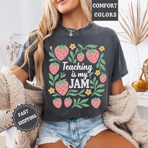 May include: Dark gray t-shirt with a strawberry and floral design. The text "Teaching is my JAM" is centered within the design. The shirt is worn with denim shorts and a cream-colored sweater.