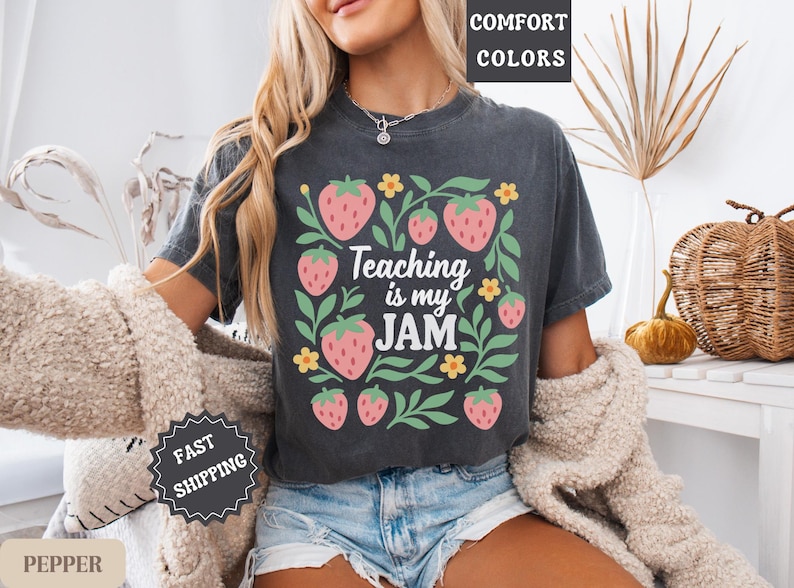 May include: Dark gray t-shirt with a strawberry and floral design. The text "Teaching is my JAM" is centered within the design. The shirt is worn with denim shorts and a cream-colored sweater.