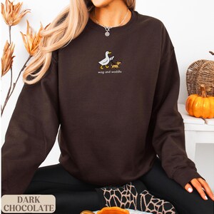 May include: Dark chocolate brown sweatshirt with the text "wag and waddle" and a graphic of a duck and dog. The sweatshirt is a long-sleeved crew neck. The words "Dark Chocolate" are printed at the bottom of the image.