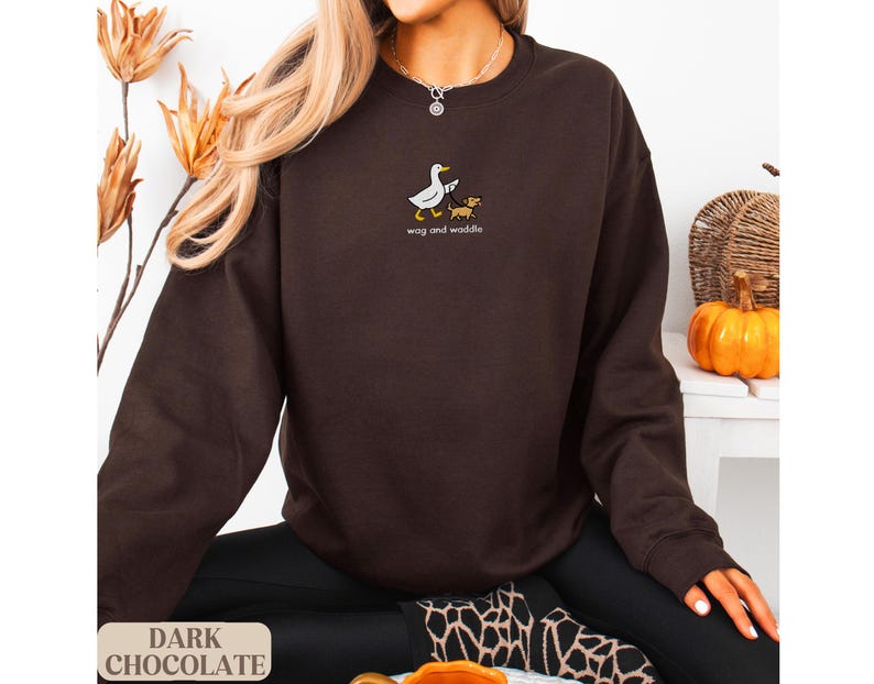 May include: Dark chocolate brown sweatshirt with the text "wag and waddle" and a graphic of a duck and dog. The sweatshirt is a long-sleeved crew neck. The words "Dark Chocolate" are printed at the bottom of the image.