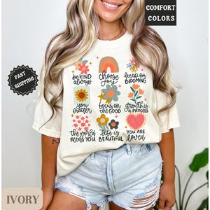 Pu&ograve; includere: Una maglietta color crema con un design floreale e affermazioni positive. Il testo sulla maglietta recita "Sii gentile sempre", "Scegli la gioia", "Continua a fiorire", "Tu conti", "Concentrati sul bene", "La crescita &egrave; un processo", "Il mondo ha bisogno di te", "La vita &egrave; bella", e "Sei amato".