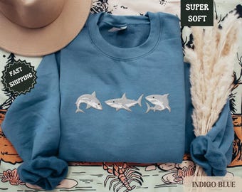 May include: Indigo blue sweatshirt with embroidered white sharks. The sweatshirt has a crew neck and long sleeves. The text "SUPER SOFT" is printed on the top right corner of the image.