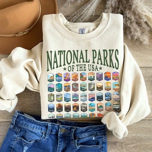 May include: A cream-colored sweatshirt with a green graphic that says "National Parks of the USA" and features a grid of colorful national park logos.