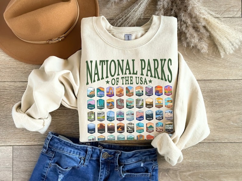 May include: A cream-colored sweatshirt with a green graphic that says "National Parks of the USA" and features a grid of colorful national park logos.