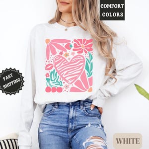 May include: White long-sleeve shirt with a pink floral and heart design. The design includes a zebra-striped heart, flowers, leaves, and cherries. The words "COMFORT COLORS" and "WHITE" are visible.