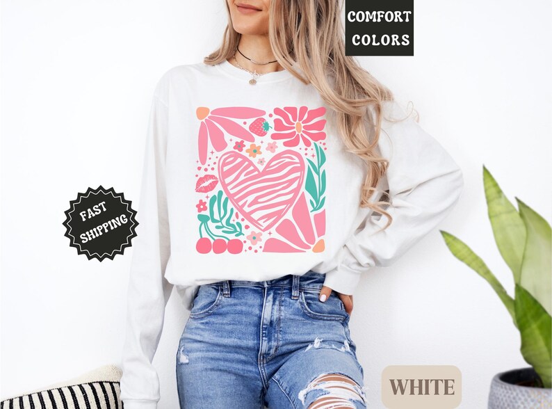 May include: White long-sleeve shirt with a pink floral and heart design. The design includes a zebra-striped heart, flowers, leaves, and cherries. The words "COMFORT COLORS" and "WHITE" are visible.