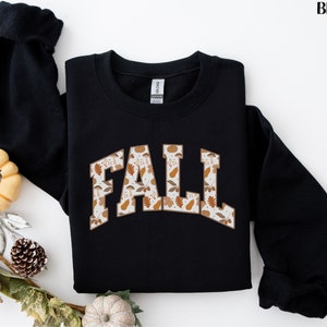 K&ouml;nnte beinhalten: Schwarzer Pullover mit einem wei&szlig;en "FALL"-Motiv mit einem Muster aus Herbstbl&auml;ttern und K&uuml;rbissen.